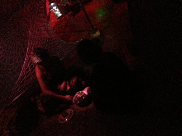 In very low light tinged with red, a woman who is a seer sits on the ground with her enchanted saucepan observing people, who crouch before her to hear her wisdoms